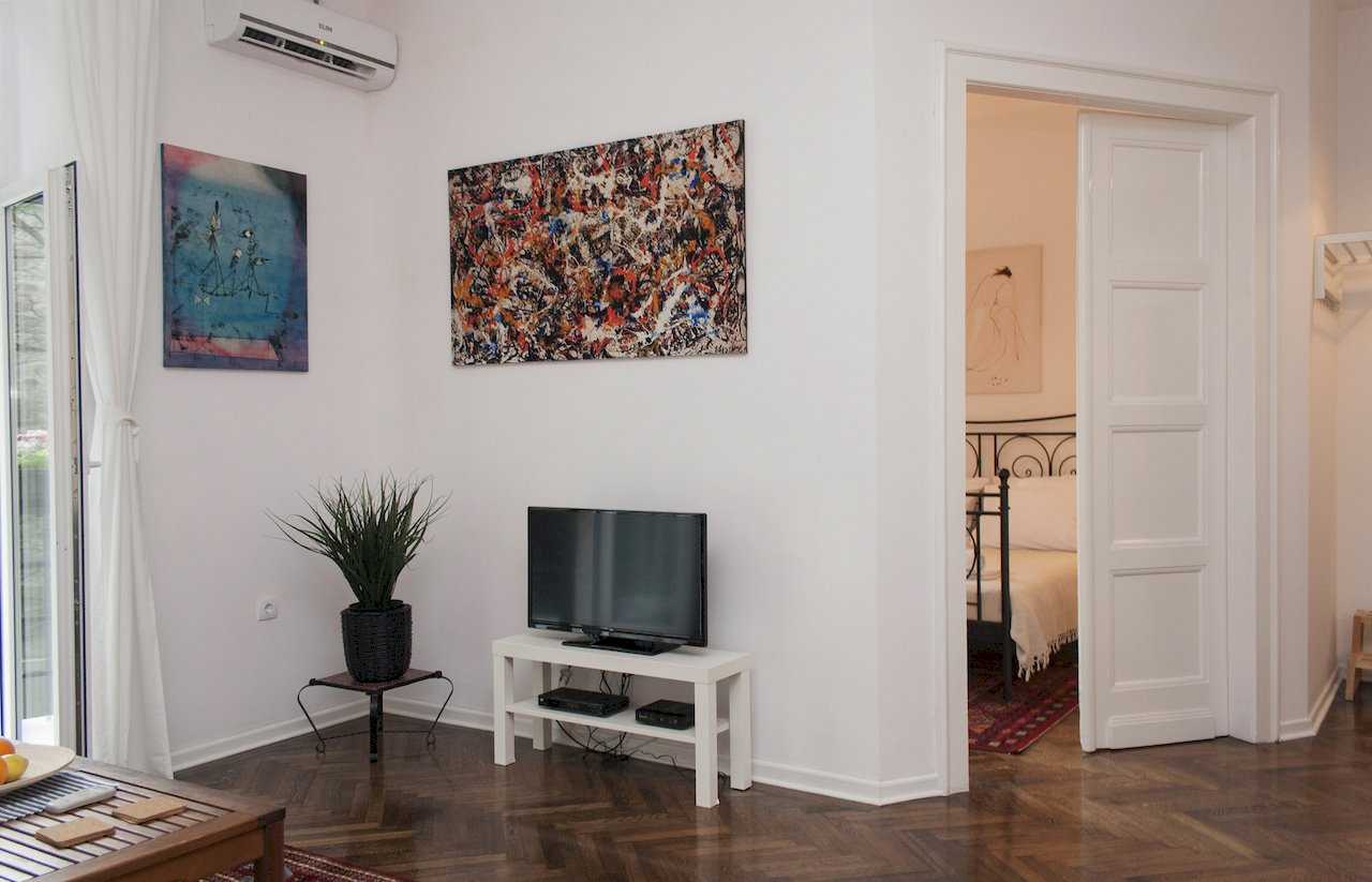 Living area with flat-screen TV on a white console, wall-mounted air conditioner, two abstract paintings (Pollock-style and Miró-style) and view through open doors into bedroom with black wrought-iron bed – Apartment Atelje 2 – City Break Apartments