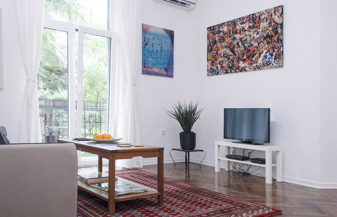 Living room with sliding balcony doors and sheer curtains, wooden coffee table holding a bowl of oranges on a red oriental rug, potted plant on a wrought-iron stand and TV on a white console – Apartment Atelje 2 – City Break Apartments