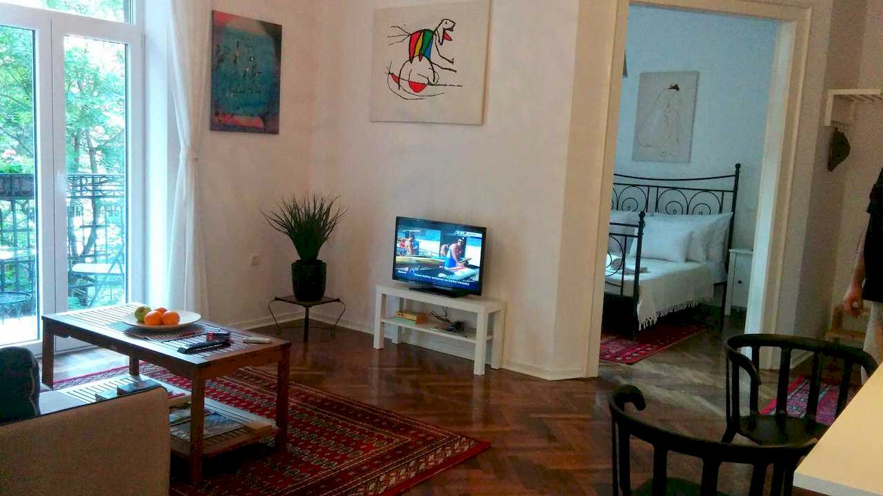 Living area with coffee table holding oranges, switched-on TV, two abstract artworks (one a colourful figurative piece) and an open doorway revealing the bedroom with black wrought-iron bed – Apartment Atelje 2 – City Break Apartments