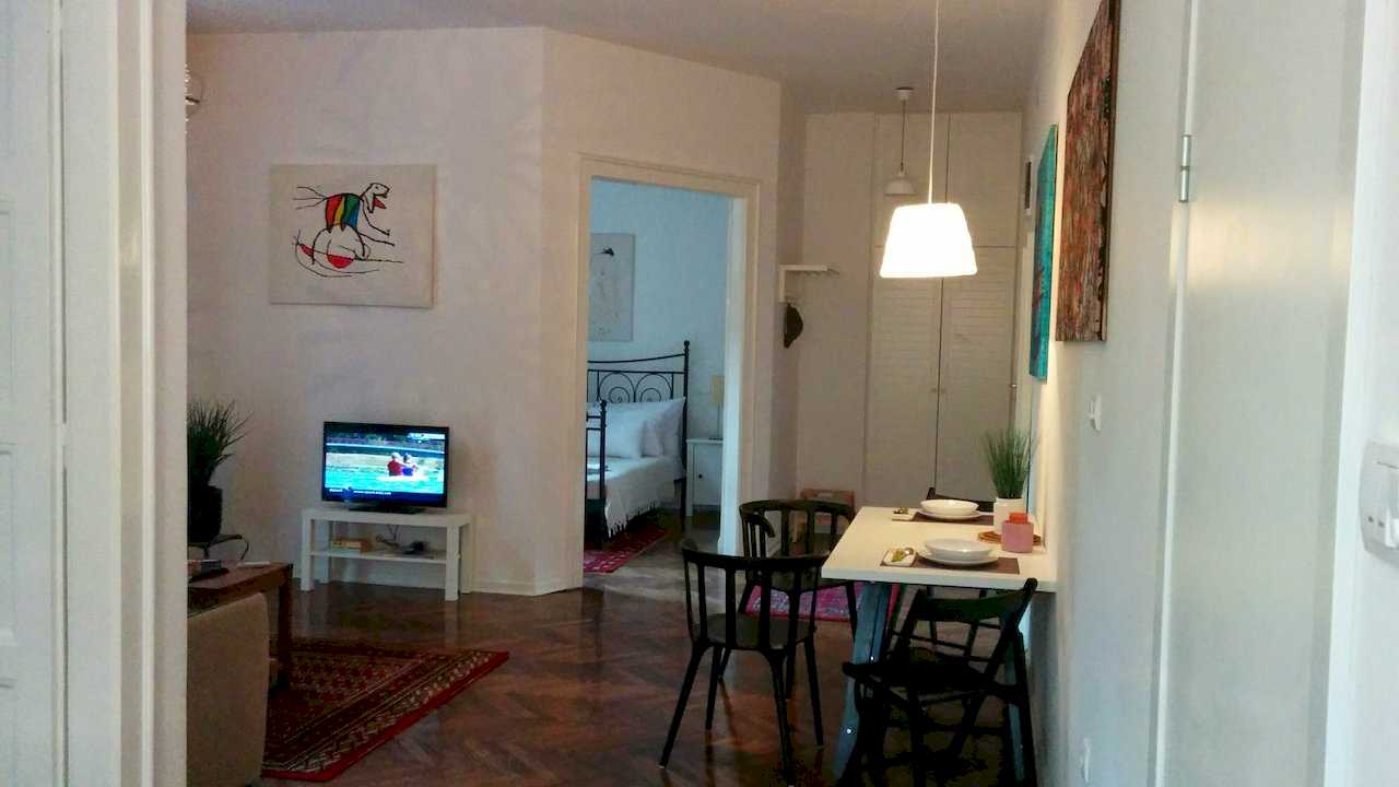 Wide view from the hallway across the living zone with TV, dining table for two with black chairs and an open door into the bedroom with wrought-iron bed – Apartment Atelje 2 – City Break Apartments