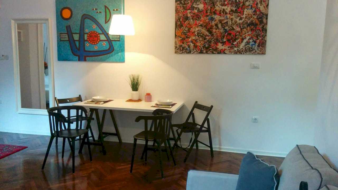 Dining nook with white table on black X-trestle legs, folding black chairs, pendant lamp above and two large abstract canvases (blue Miró-style and red-toned Pollock-style) on the white wall – Apartment Atelje 2 – City Break Apartments
