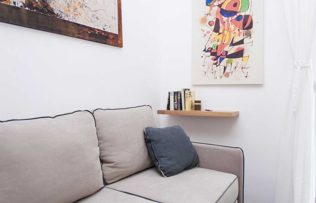 Beige sofa corner with blue cushion, floating wooden shelf with books and two framed abstract artworks (Miró-style and dark brown) on a white wall – Apartment Atelje 2 – City Break Apartments