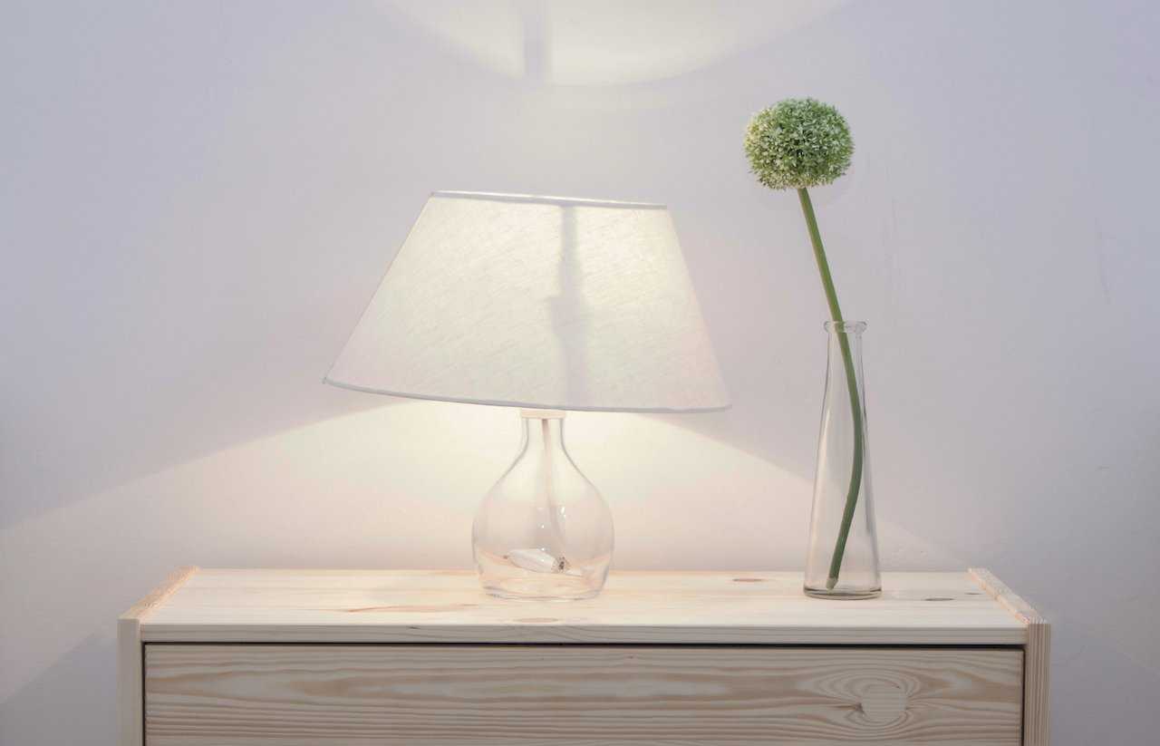 Detail of light pinewood dresser with glass-base lamp and white shade beside a slim glass vase holding a green allium bloom – Apartment Atelje 2 – City Break Apartments