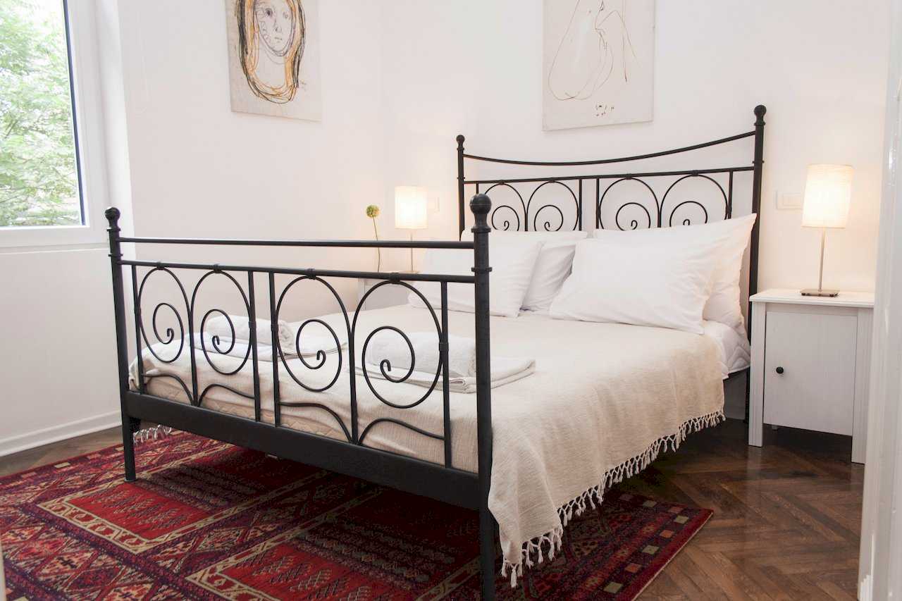 Bedroom with black wrought-iron double bed (scroll motifs), burgundy oriental rug, two white nightstands with table lamps and minimalist sketch portraits above the headboard – Apartment Atelje 2 – City Break Apartments