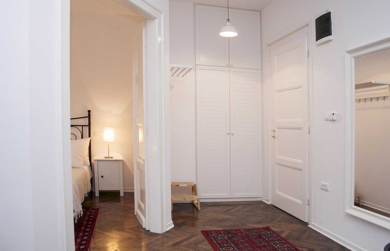 Hallway with floor-to-ceiling white wardrobe, white entrance door with intercom, small wooden step-stool and view through open doors into the bedroom with wrought-iron bed – Apartment Atelje 2 – City Break Apartments