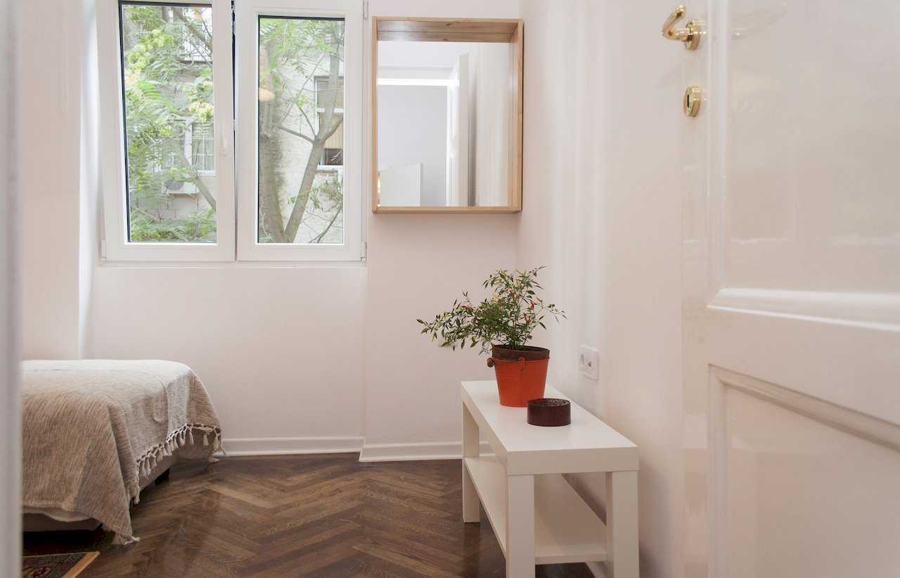 Bedroom corner with double window overlooking trees and a neighbouring building, framed wooden mirror, small white bench with orange potted plant and edge of the bed in the foreground – Apartment Atelje 2 – City Break Apartments