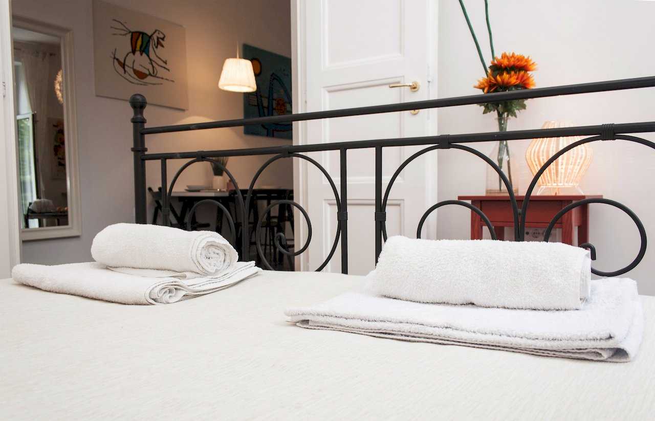 Double bed with rolled white towels, black wrought-iron headboard and view through open doors into the dining area and a small red vintage chest holding a vase of orange gerbera daisies – Apartment Atelje 2 – City Break Apartments