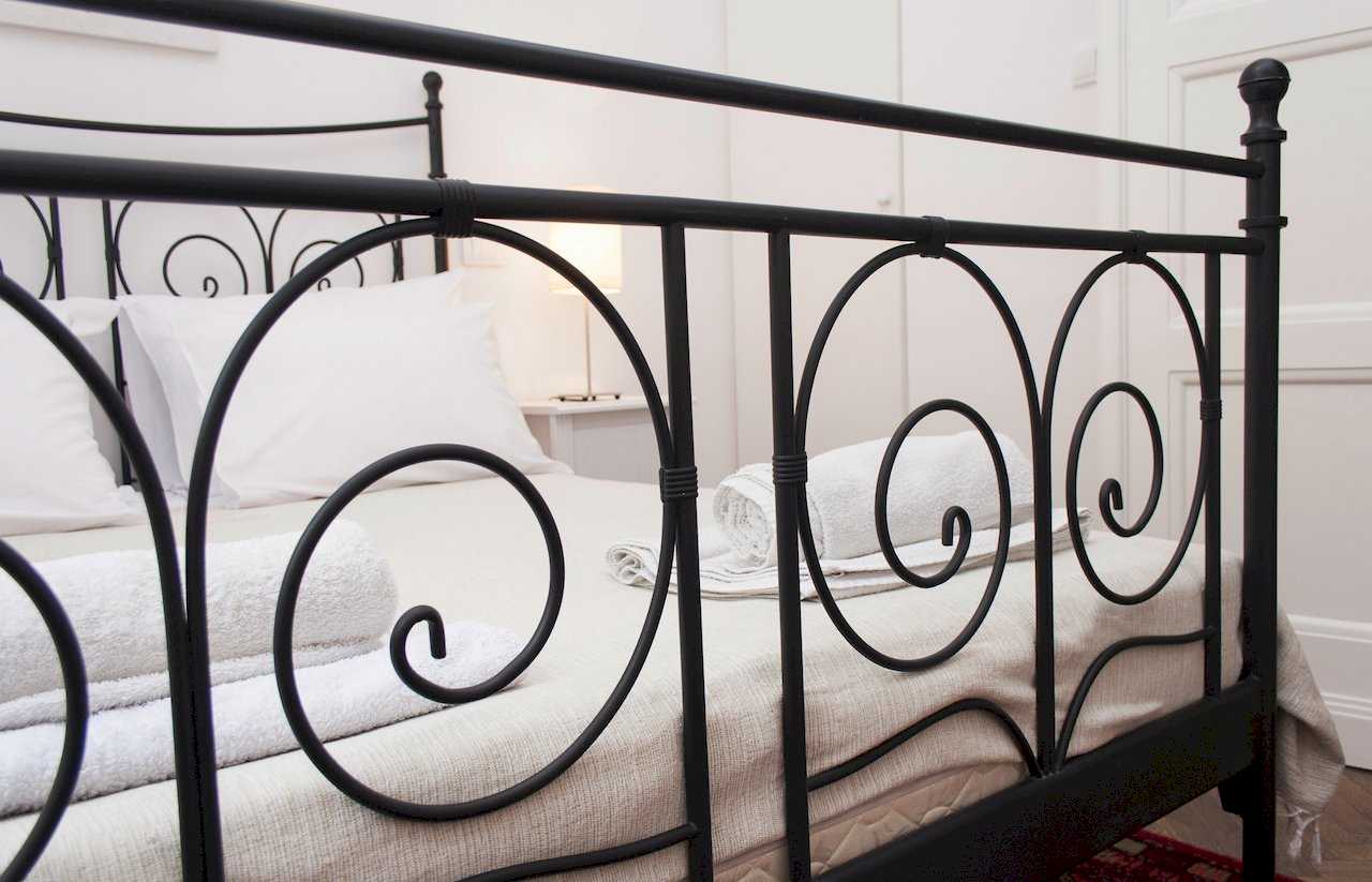 Close-up of black wrought-iron footboard with scroll motifs, white linens, rolled white towels on the bed and a glowing nightstand lamp in the background – Apartment Atelje 2 – City Break Apartments