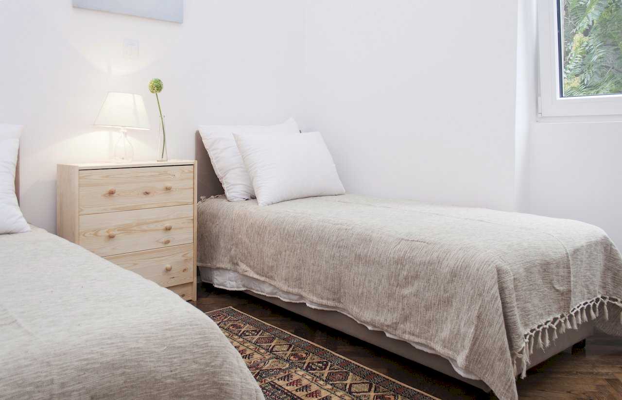 Second bedroom with two single beds, pinewood three-drawer chest with lamp and vase between them, patterned runner rug and window with green view – Apartment Atelje 2 – City Break Apartments