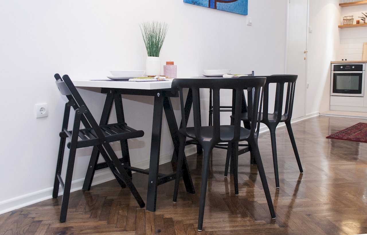 Dining table with white top on black X-trestle legs and four black chairs (folding and Starck-style), with white kitchen and built-in oven beyond – Apartment Atelje 2 – City Break Apartments