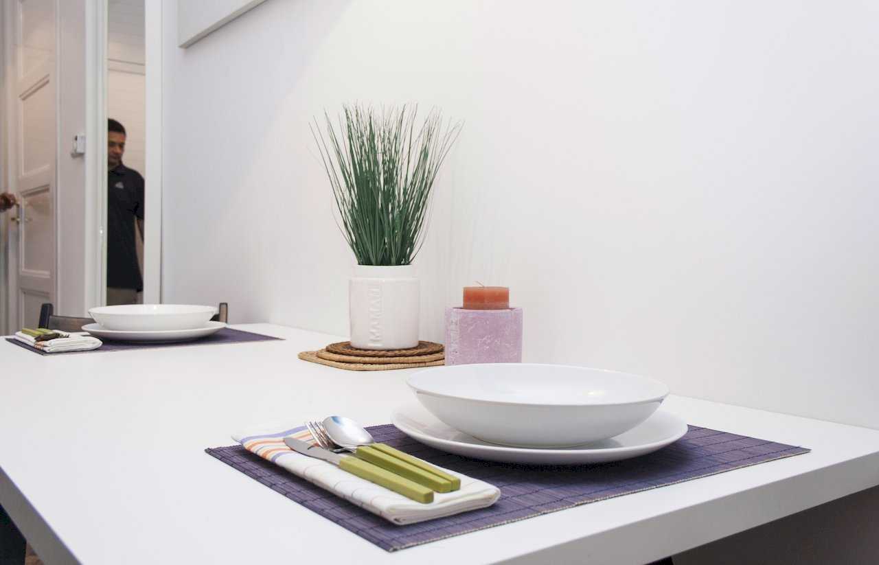Close-up of dining table for two – purple bamboo placemats, white plates, cutlery with lime-green handles, vase with grass and a small pink candle – Apartment Atelje 2 – City Break Apartments