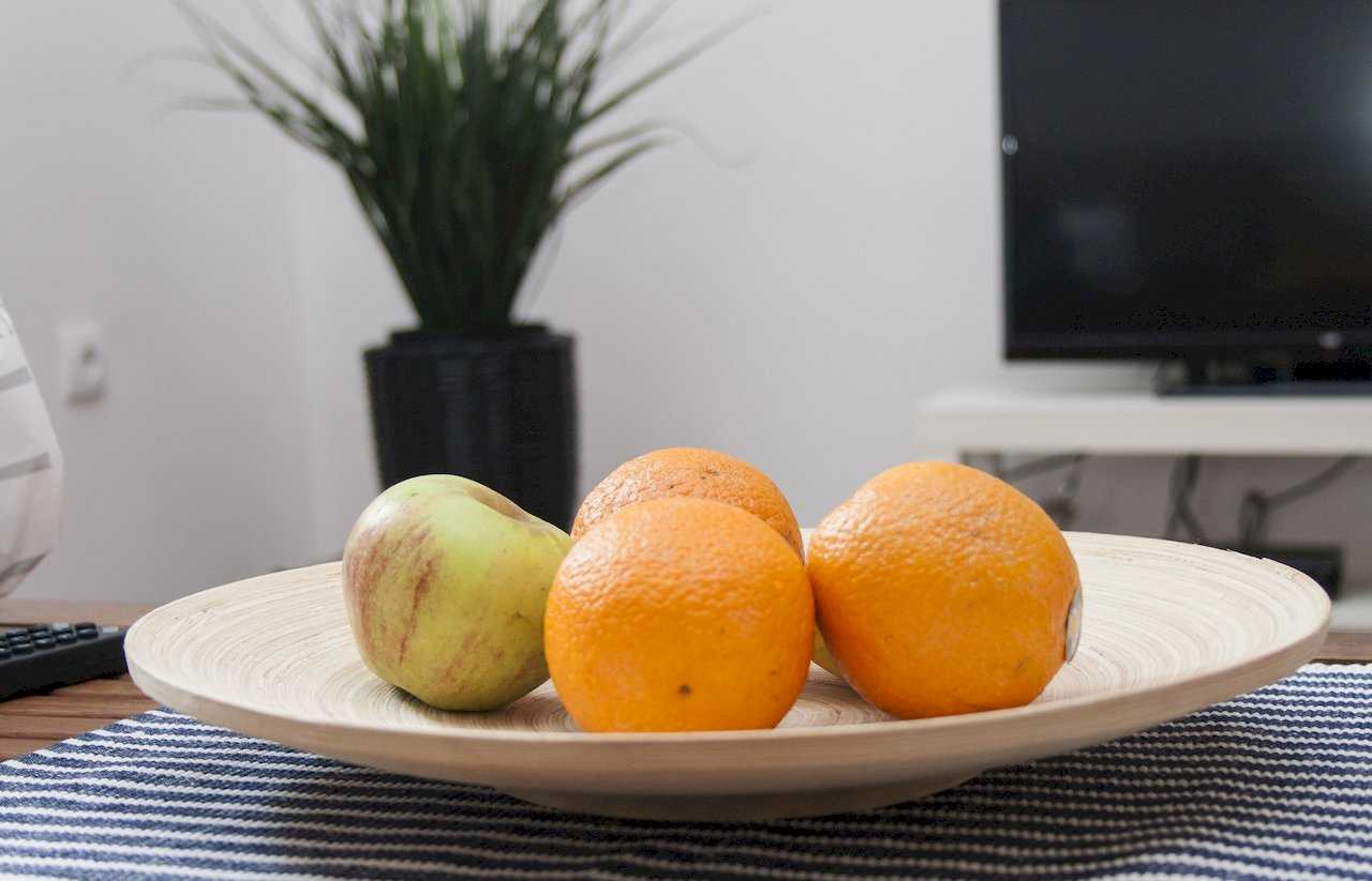 Wooden bowl with oranges and an apple on a coffee table with blue-and-white striped runner, plant and white TV console softly out of focus in the background – Apartment Atelje 2 – City Break Apartments