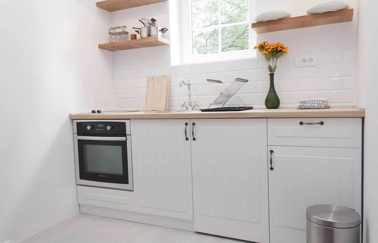 Kitchen with white cabinetry, wooden countertop, built-in Beko oven, stainless sink with classic chrome tap, open wooden shelves by the window and sunflowers in a green vase – Apartment Atelje 2 – City Break Apartments