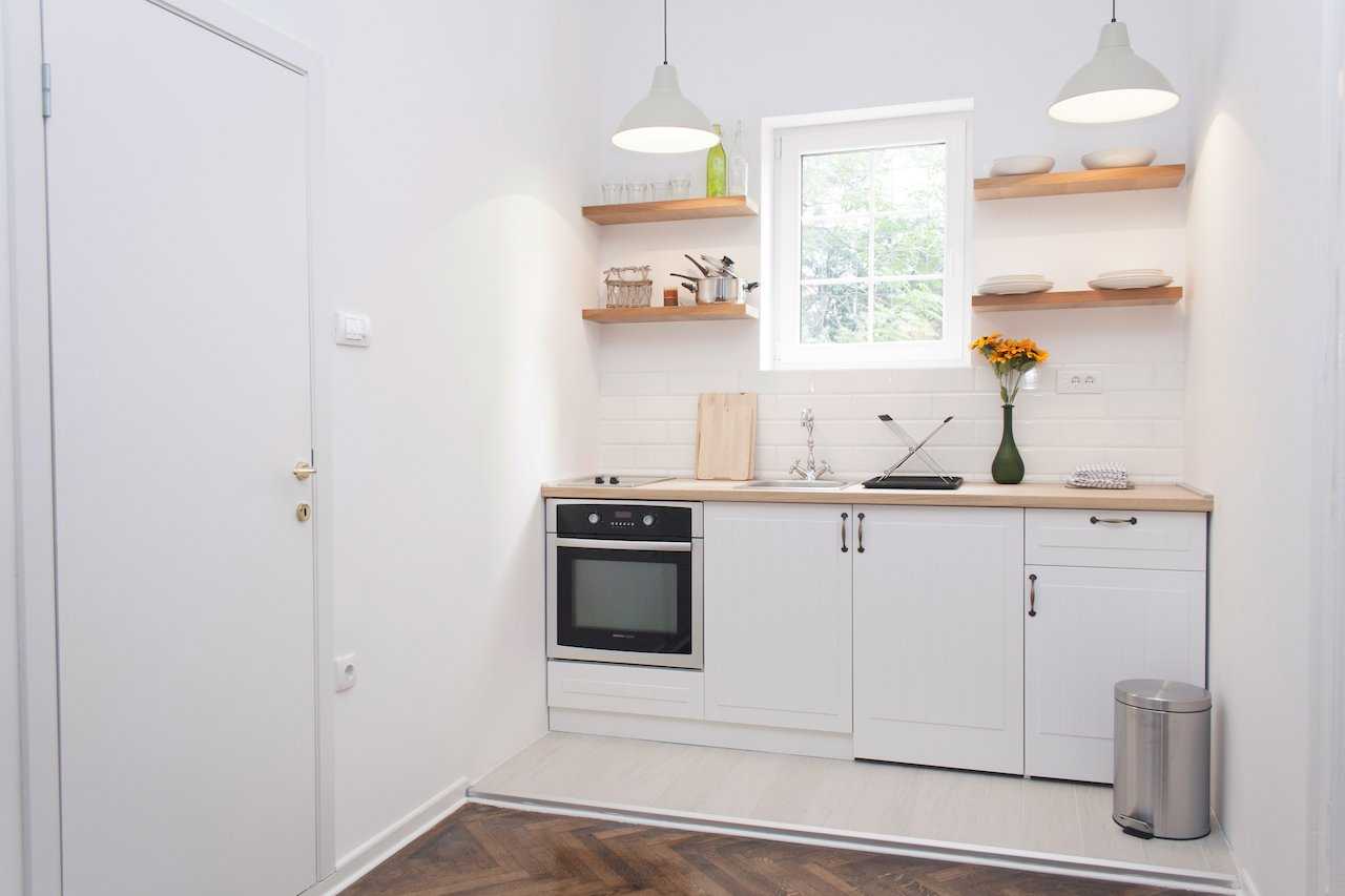 Wider view of the kitchen with twin white pendant lights, open wooden shelves, built-in oven and hob, classic chrome tap and sunflowers on the counter – seen from the hallway – Apartment Atelje 2 – City Break Apartments