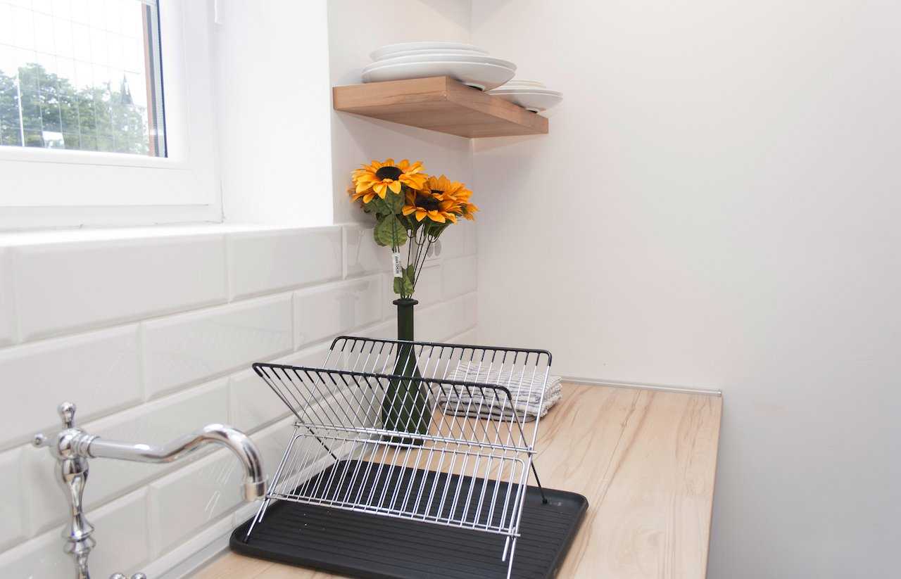 Kitchen corner by the window – wire dish rack on the wood counter, vase with fresh sunflowers and floating wooden shelf with white plates against a subway-tile splashback – Apartment Atelje 2 – City Break Apartments