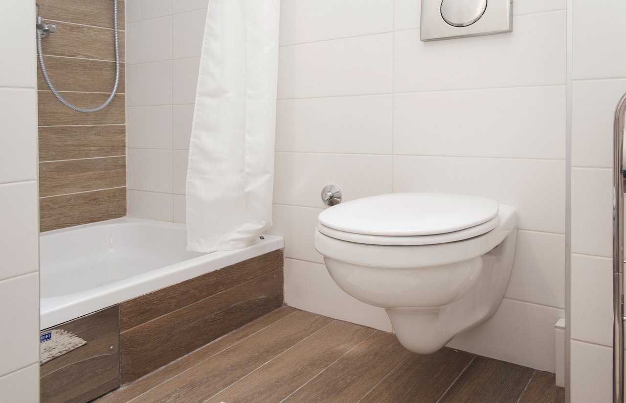 Bathroom with wall-hung white toilet, bathtub with white shower curtain and handheld shower, plus wood-look accent tiling around the tub – Apartment Atelje 2 – City Break Apartments