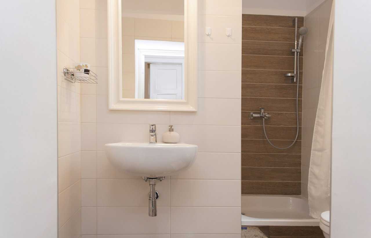 Bathroom from another angle – wall-mounted white sink with chrome tap, large mirror in a white frame and shower zone with wood-look tiles and wall-mounted shower mixer – Apartment Atelje 2 – City Break Apartments