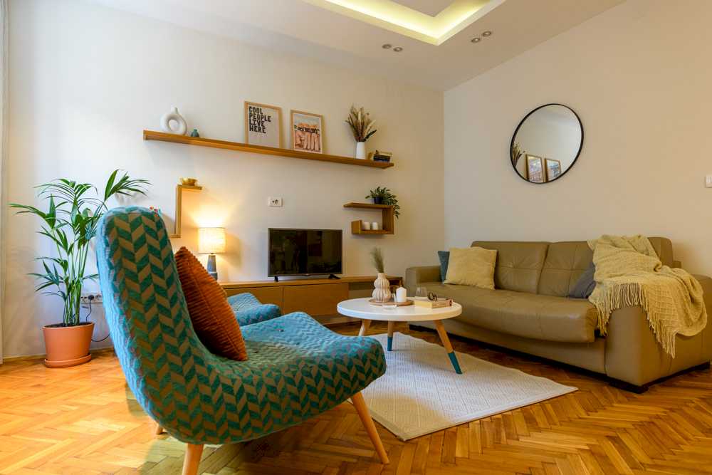 Širi pogled na dnevni boravak sa bež kožnim dvosedom, jednom tirkiznom mid-century foteljom, palmom u terakota saksiji, drvenim zidnim policama i velikim okruglim ogledalom u crnom ramu – Apartman Design – City Break Apartments