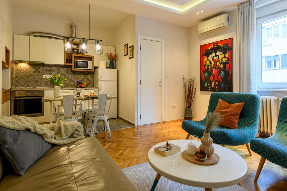 Open-plan dnevna soba sa kuhinjom – ugradna rerna i mikrotalasna, sivi cementni patchwork backsplash, mermerni trpezarijski sto sa belim Tolix stolicama i šarena slika tri afričke žene sa šeširima na zidu pored klima uređaja – Apartman Design – City Break Apartments