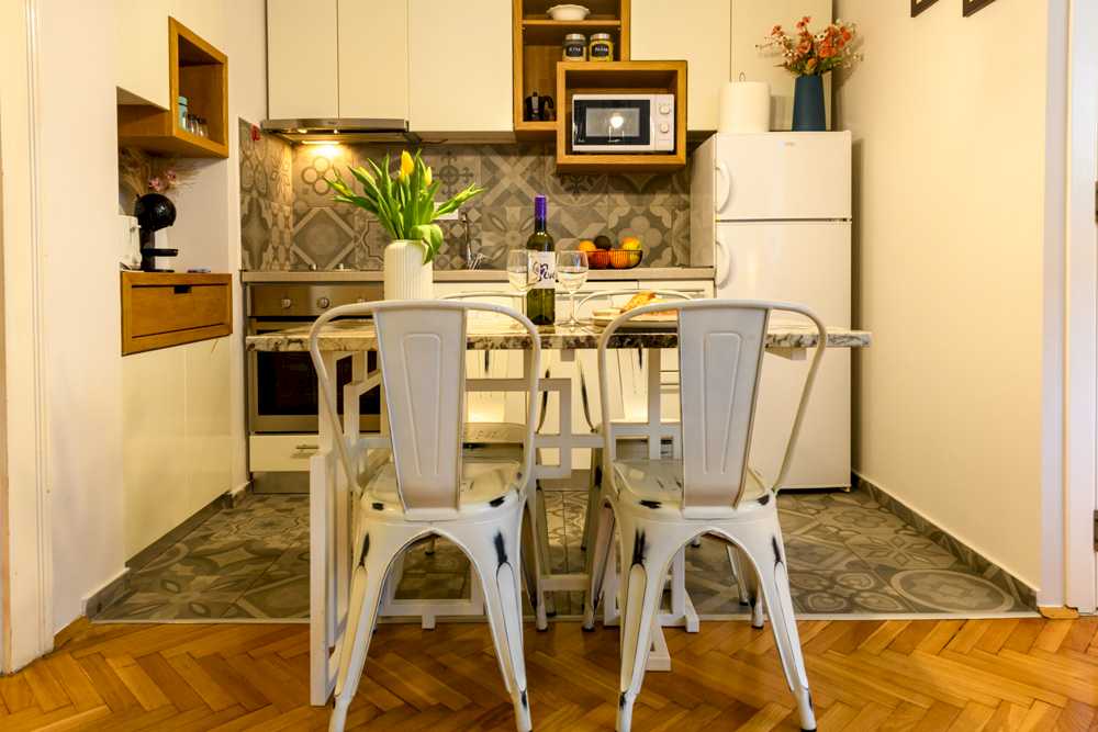 Trpezarijski sto sa žutim tulipanima u beloj vazi i flašom vina, kuhinja sa patchwork cementnim pločicama, ugradnom rernom, mikrotalasnom na drvenom rafu i belim frižiderom – Apartman Design – City Break Apartments