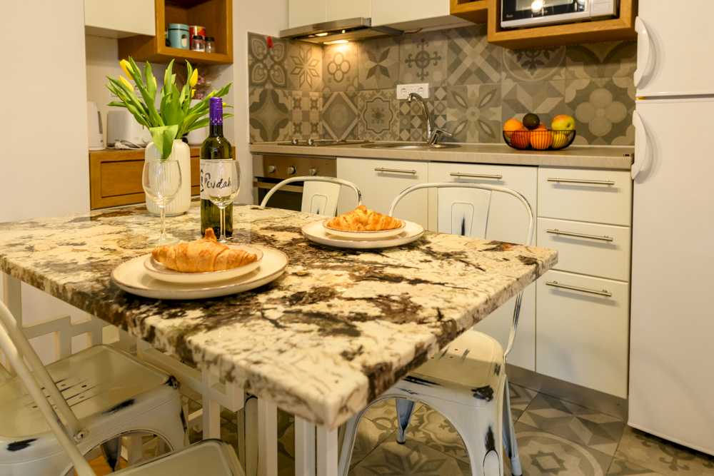 Krupni plan mermernog trpezarijskog stola – beli tanjiri sa kroasanima, vaza sa žutim tulipanima i flaša 'Rudak' vina, u pozadini cementni patchwork backsplash i bele kuhinjske ladice – Apartman Design – City Break Apartments