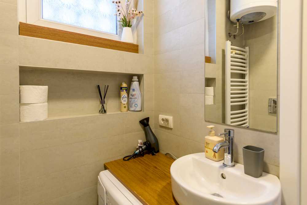 Kupatilo sa belim ovalnim umivaonikom i hrom slavinom, ogledalom, niše-policom sa toaletnim papirom i diffuser-om, bojlerom u uglu i drvenom pločom preko mašine za veš sa fenom – Apartman Design – City Break Apartments