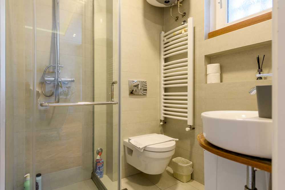 Kupatilo sa kliznom staklenom tuš kabinom, wall-hung belim WC-om sa složenim peškirom, ovalnim umivaonikom na drvenoj polici i belom električnom sušilicom za peškire – Apartman Design – City Break Apartments