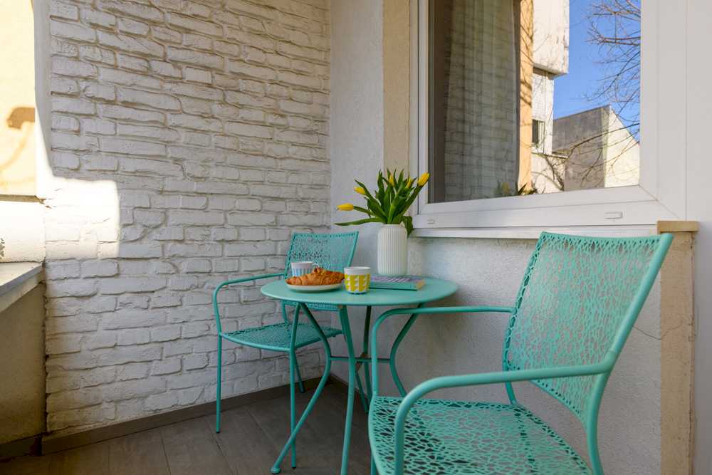 Mala terasa sa zidom od kreč-bele cigle, mint okruglim metalnim stolom i dve mint stolice u lace dezenu, vazom sa žutim tulipanima i poslužavnikom sa kroasanima i kafom – Apartman Design – City Break Apartments