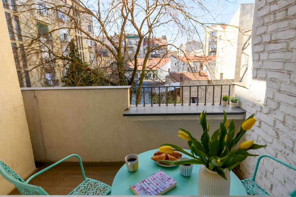 Pogled sa terase preko kovano-gvozdene ograde na unutrašnje dvorište sa starim beogradskim krovovima i golim krošnjama drveća; u prvom planu mint sto sa žutim tulipanima i kroasanima – Apartman Design – City Break Apartments