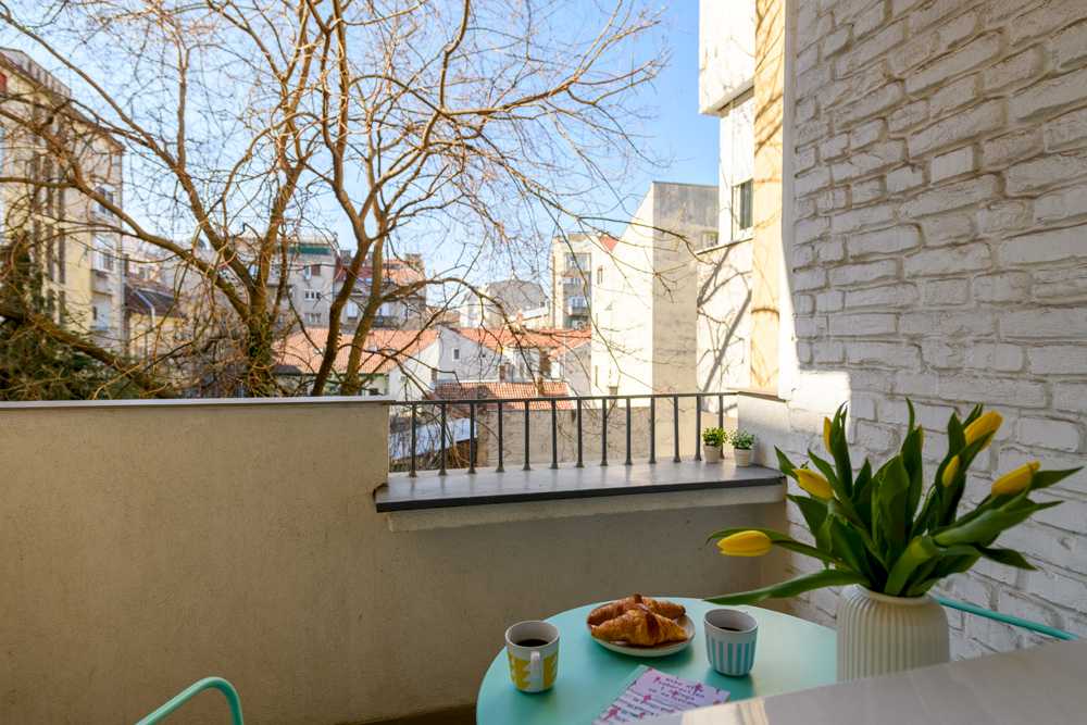 Pogled sa terase na okolne zgrade i krošnje, kovano-gvozdena ograda, mint sto sa žutim tulipanima u beloj vazi, kafom i kroasanima u prvom planu – Apartman Design – City Break Apartments