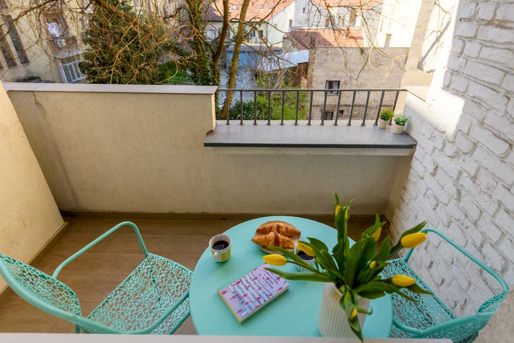 Pogled odozgo na terasu – mint okrugli sto sa žutim tulipanima, mint lace stolice, knjiga, kafa i kroasani, kovano-gvozdena ograda i pogled na krovove – Apartman Design – City Break Apartments