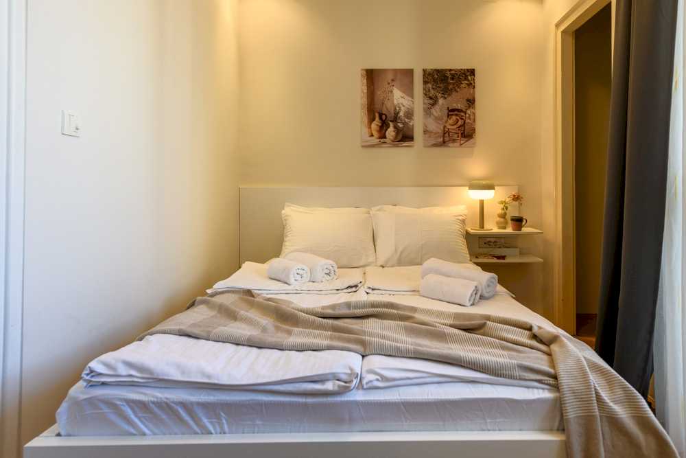Spavaća soba sa bračnim krevetom, dva para belih peškira u rolnama, mushroom-lampa na beloj zidnoj polici sa malom vazom i šoljom kafe i dve uokvirene boho fotografije iznad uzglavlja – Apartman Design – City Break Apartments