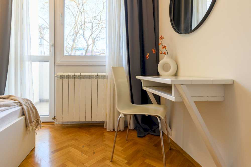 Radni kutak u spavaćoj sobi – bela zidna radna ploča sa drvenom dijagonalnom nogom, bela moderna stolica, donut-vaza sa narandžastim grančicama i pogled na balkonska vrata – Apartman Design – City Break Apartments
