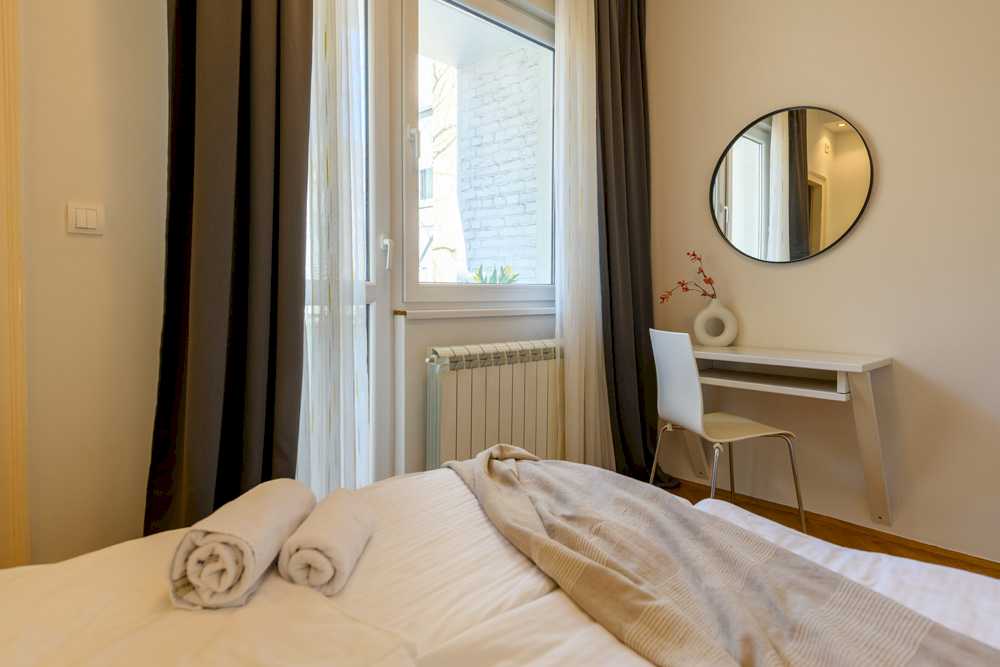 Pogled iz kreveta na radni kutak – bela radna ploča sa donut-vazom, bela moderna stolica, crno okruglo ogledalo i prozor sa pogledom na susednu zgradu – Apartman Design – City Break Apartments