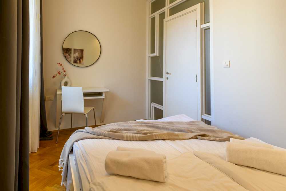 Pogled iz spavaće sobe ka francuskim vratima sa zelenim staklenim okvirima i radnom kutku sa belim stolom, donut-vazom i okruglim crnim ogledalom – Apartman Design – City Break Apartments
