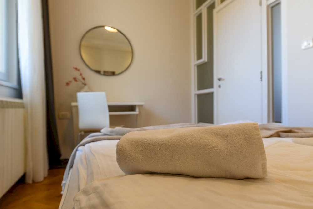 Detalj spavaće sobe – sklopljen bež peškir u prvom planu, u izmaglici radni sto sa donut-vazom, ogledalo i francuska vrata – Apartman Design – City Break Apartments