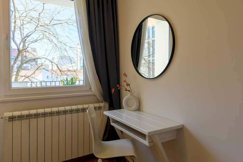 Radni kutak uz prozor sa pogledom na okolne zgrade i gole krošnje, bela radna ploča sa donut-vazom sa narandžastim grančicama, bela stolica i crno okruglo ogledalo iznad – Apartman Design – City Break Apartments