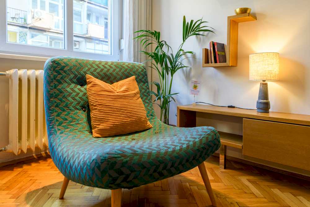 Detalj sedeće zone – tirkizna mid-century fotelja sa narandžastim jastukom, palma u terakota saksiji, drvena TV komoda sa lampom i zidna geometrijska polica sa knjigama – Apartman Design – City Break Apartments
