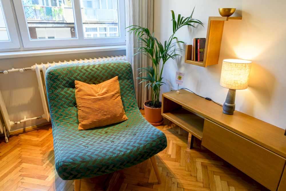 Tirkizna mid-century fotelja sa narandžastim jastukom pored palme u terakota saksiji, drvena TV komoda sa lampom i zidnom geometrijskom policom sa knjigama, prozor levo – Apartman Design – City Break Apartments