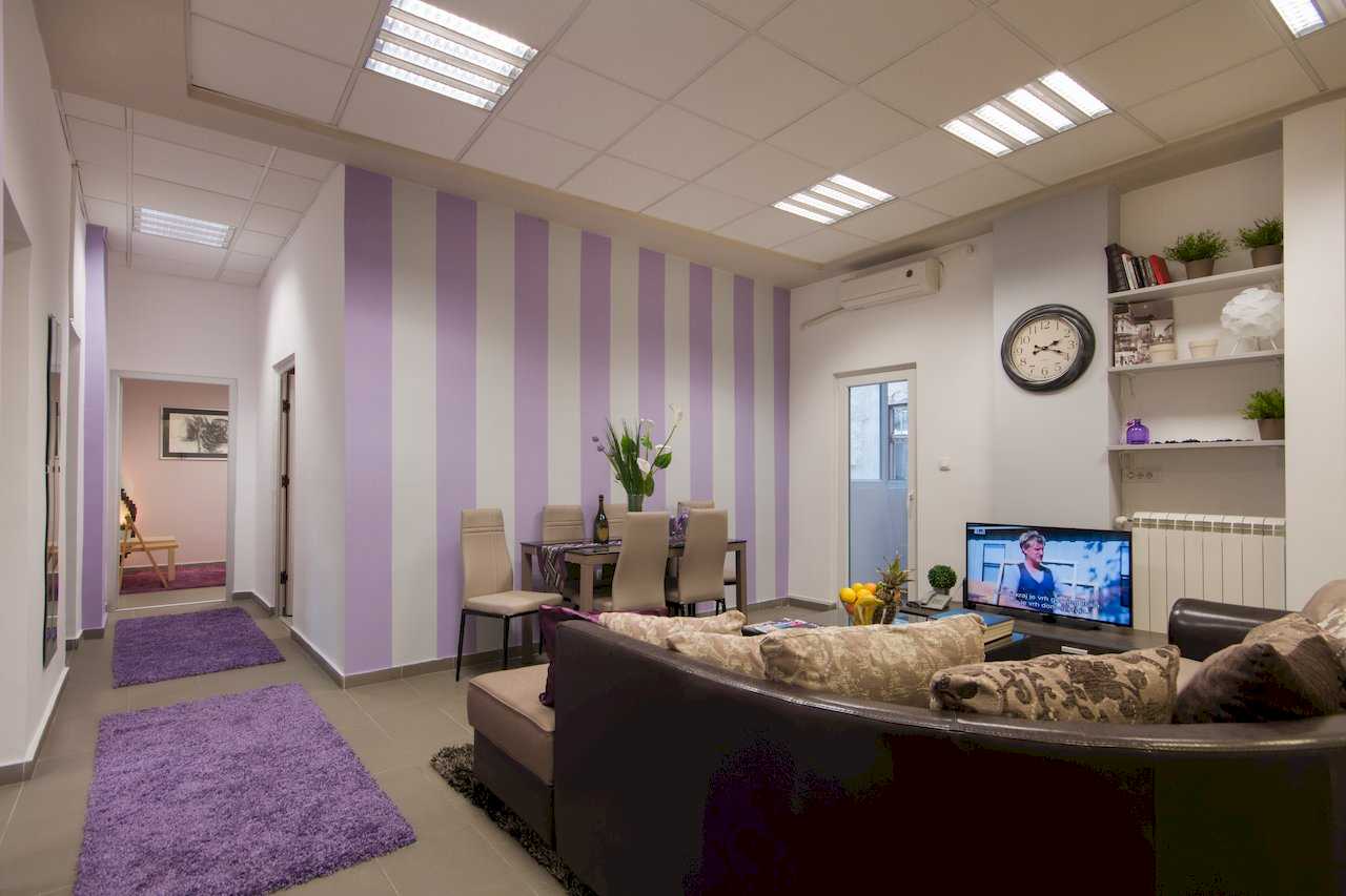 Living room with sectional sofa, TV, round wall clock, and built-in shelves; dining area with striped accent wall – Apartment Klio – City Break Apartments