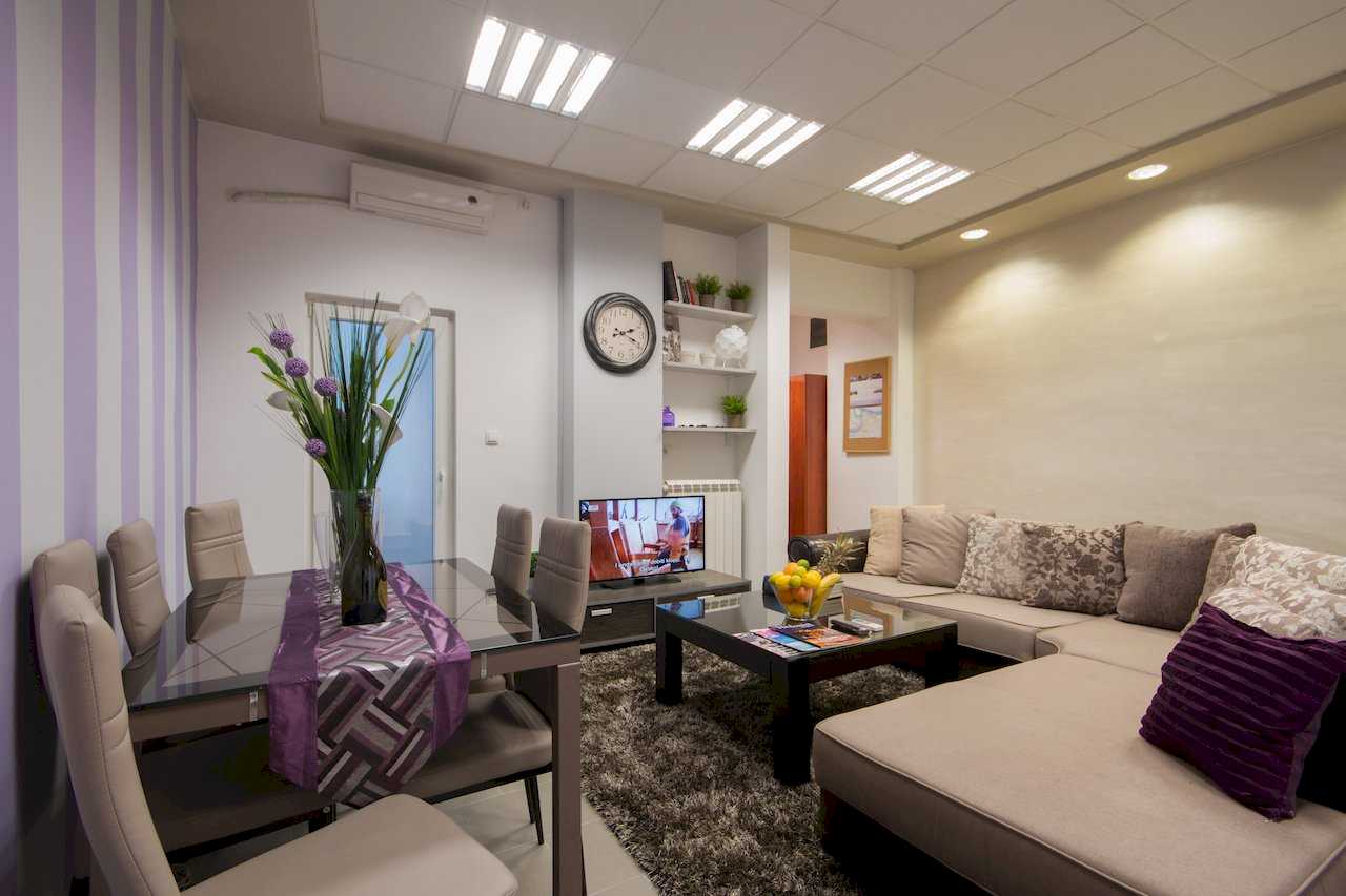 Open-plan living and dining with beige sectional, glass dining table for six, purple-and-white striped accent wall at the dining area, and a large round wall clock – Apartment Klio – City Break Apartments