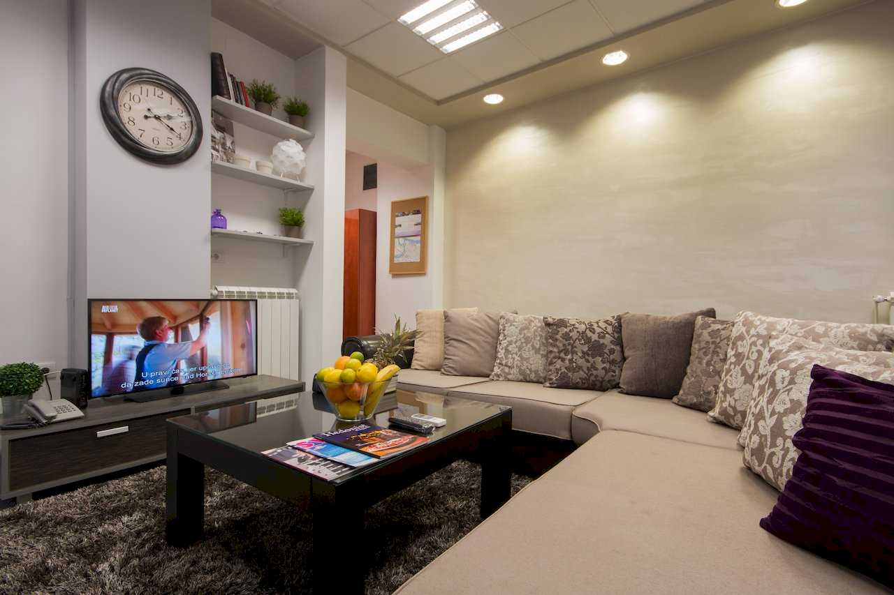 Living area with beige sectional, glass coffee table with fruit, TV console, and large wall clock – Apartment Klio – City Break Apartments