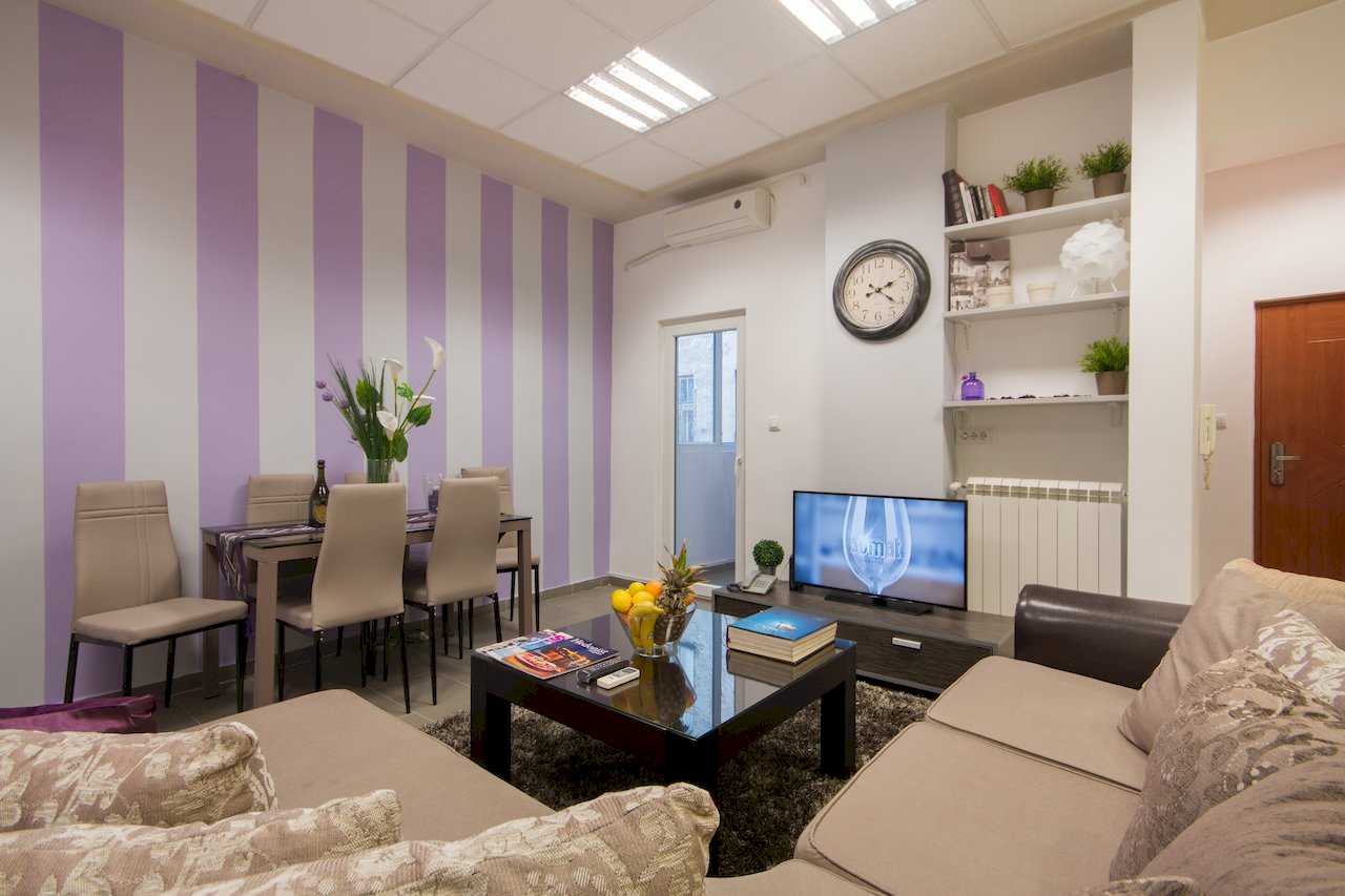 Living and dining with glass table and floral centerpiece, brown sectional, coffee table with fruit, and TV area against purple-and-white stripes – Apartment Klio – City Break Apartments
