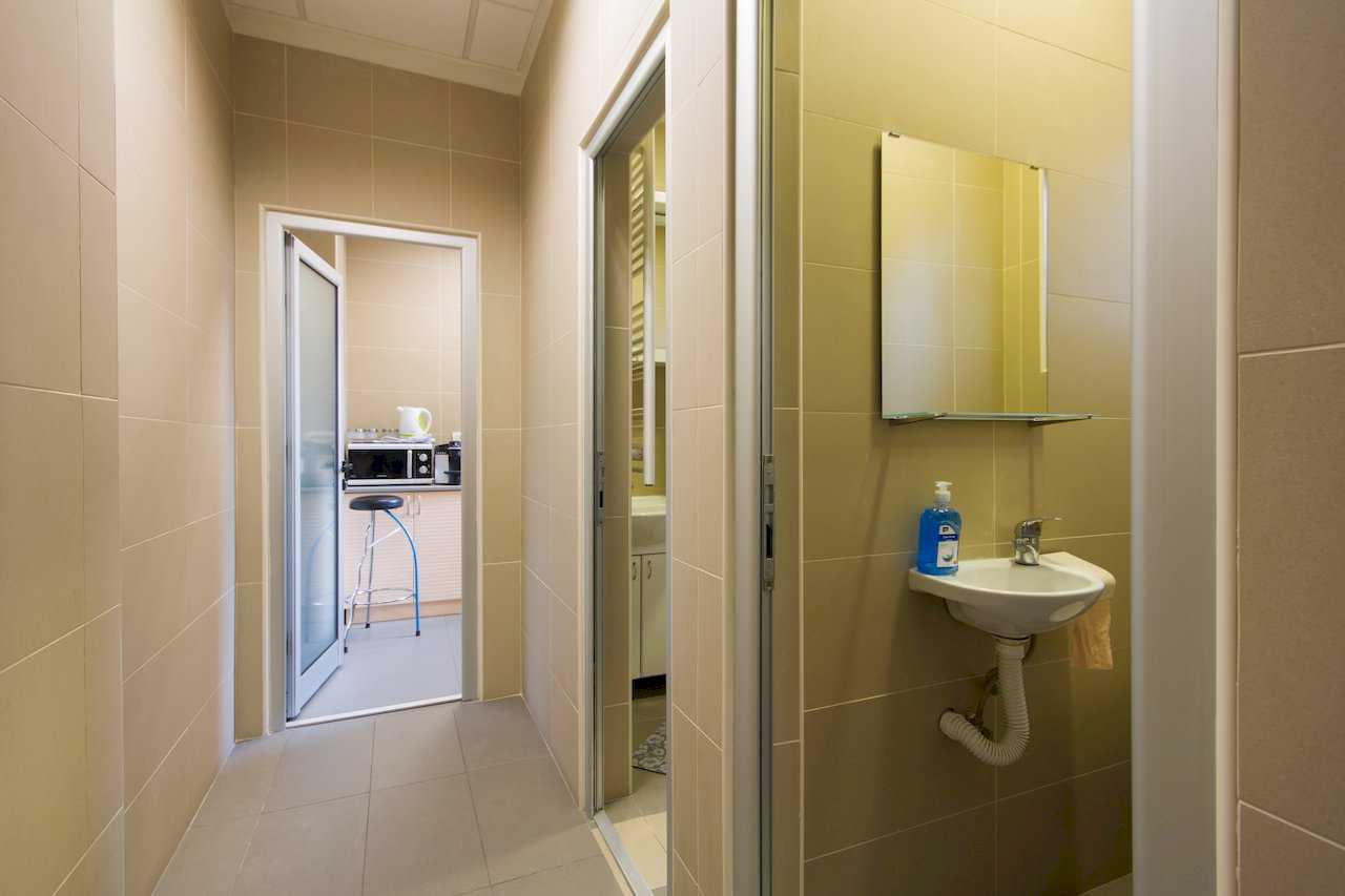 Hallway with a compact wall-mounted sink and mirror in the foreground, view into a bathroom with vanity and ladder towel radiator and into the kitchen with a bar stool – Apartment Klio – City Break Apartments