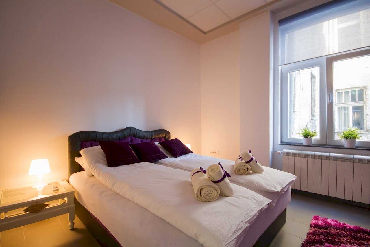 Bedroom with double bed, black upholstered headboard and deep purple pillows, rolled white towels with purple ribbons, and a large window with roller blind – Apartment Klio – City Break Apartments
