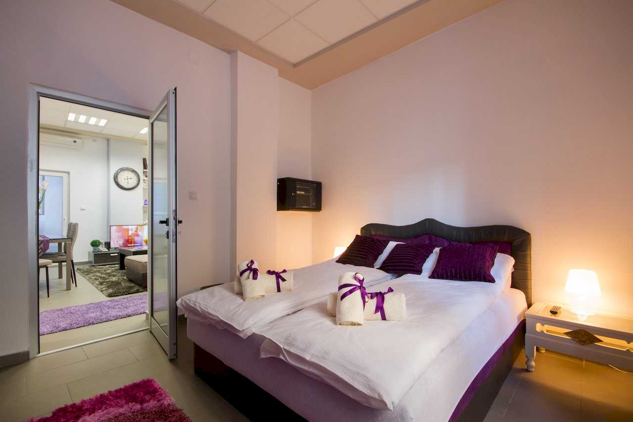 Bedroom with double bed and view through open doors into the living and dining area with TV and a purple rug – Apartment Klio – City Break Apartments