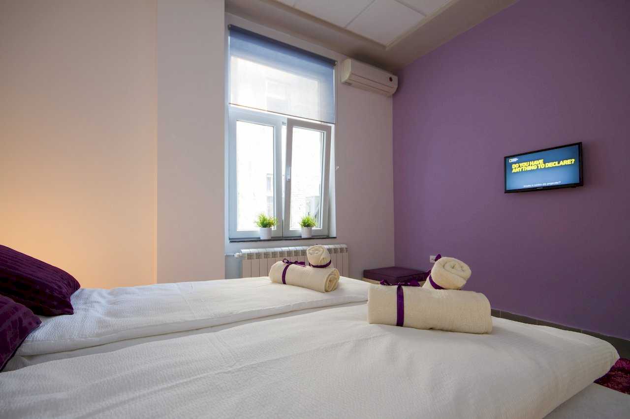 Bedroom with two joined single beds, purple accent wall, wall-mounted TV, AC, and plants on the sill – Apartment Klio – City Break Apartments