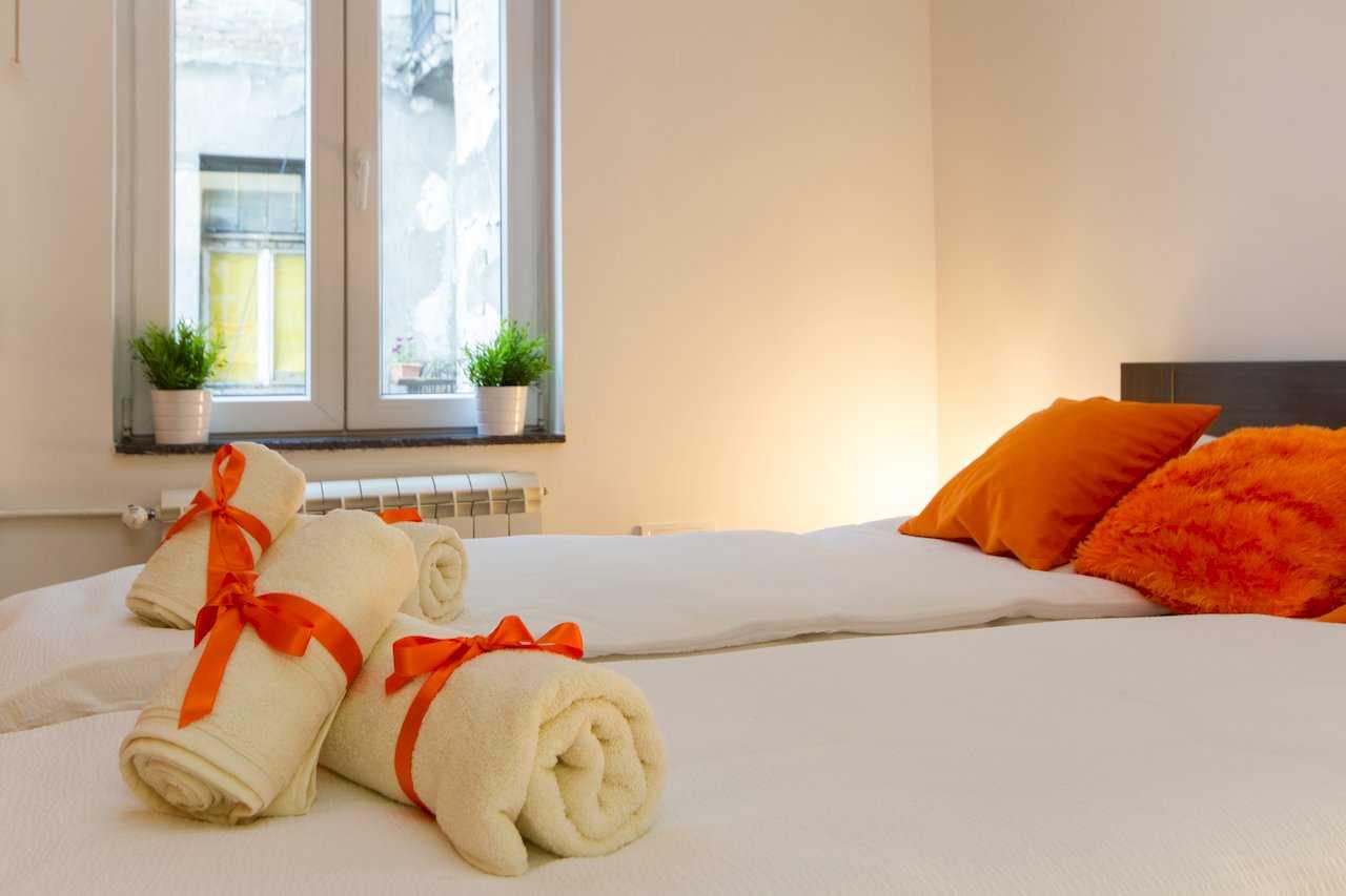 Close-up of rolled white towels with orange ribbons on the bed and plants on the windowsill – Apartment Klio – City Break Apartments