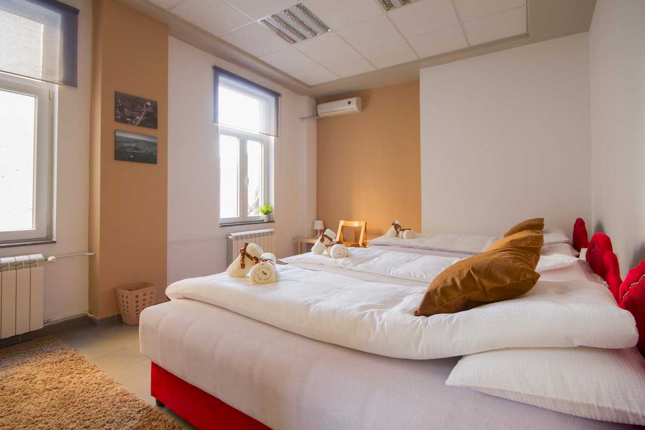 Bedroom with three single beds with red bases, white bedding, rolled towels with brown ribbons, two windows with blinds, and small framed pictures – Apartment Klio – City Break Apartments
