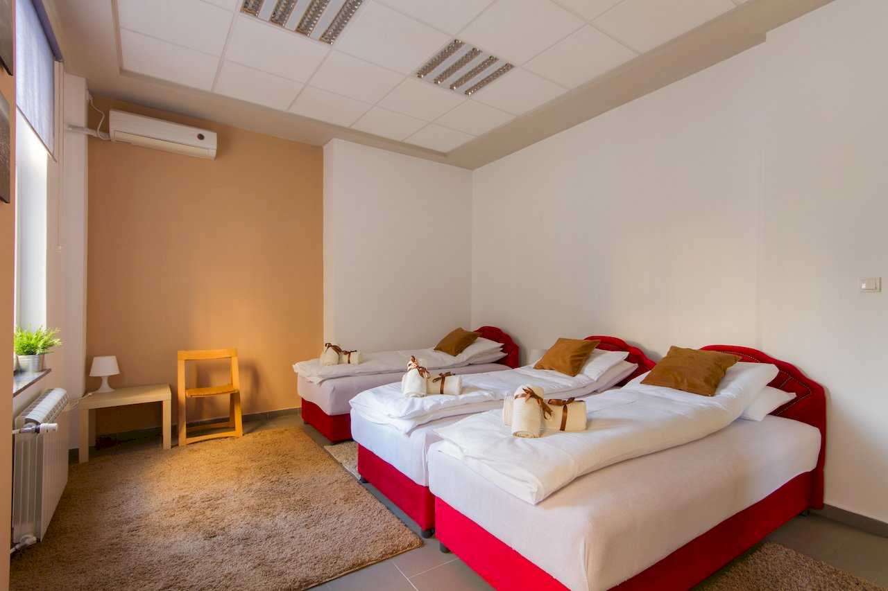 Bedroom with three single beds in a row with red bases, white linens, brown accent pillows, rolled towels with brown ribbons, beige accent wall, rug, side table with lamp, and air conditioner – Apartment Klio – City Break Apartments
